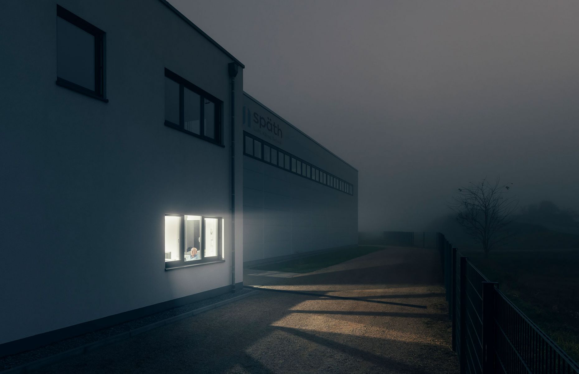 Blick von Außen auf das Gebäude bei Nebel Ein Mitarbeiter sitzt noch im erleuchteten Büro
